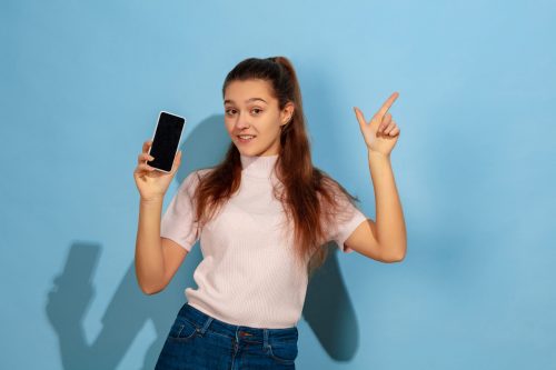 Showing phone screen, pointing up. Caucasian teen girl's portrait on blue background. Beautiful model in casual wear. Concept of human emotions, facial expression, sales, ad. Copyspace. Looks happy.
